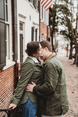 A couple wearing matching green jackets share an intimate moment on a brick sidewalk in front of historic buildings.