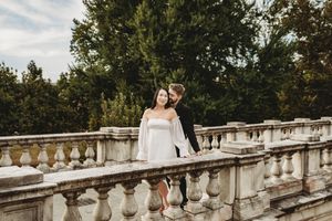 Couple in wedding attire posing on ornate stone balustrade with lush green trees in background.