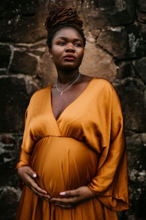 Artistic maternity portrait featuring flowing mustard dress against stone wall backdrop.