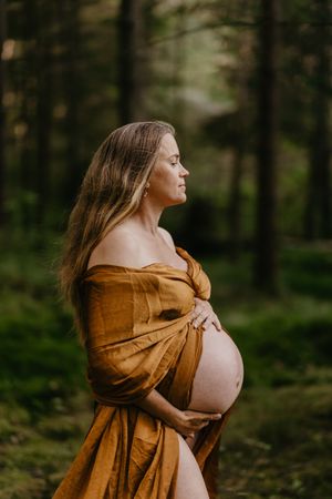A maternity photo series in a forest shows an expectant mother in a flowing brown dress embracing her baby bump.