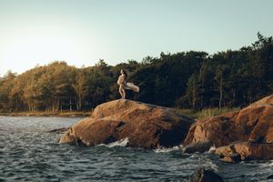 A person performs a backflip off a rocky cliff into the ocean during sunset, creating a dramatic silhouette against the sky.