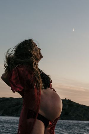 Silhouette of pregnant woman in swimwear standing against sunset sky with crescent moon over water.
