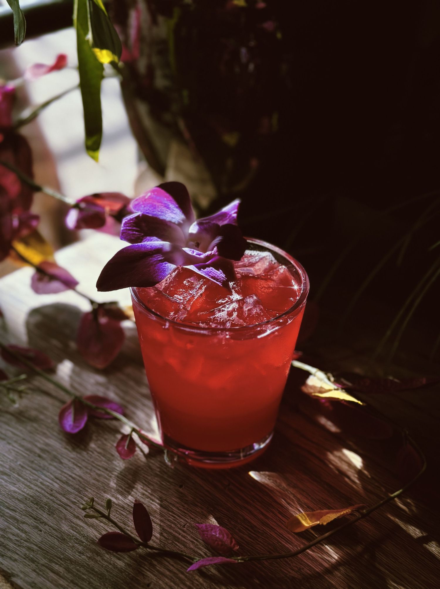 A red cocktail garnished with purple orchid flowers on a wooden surface in moody lighting.