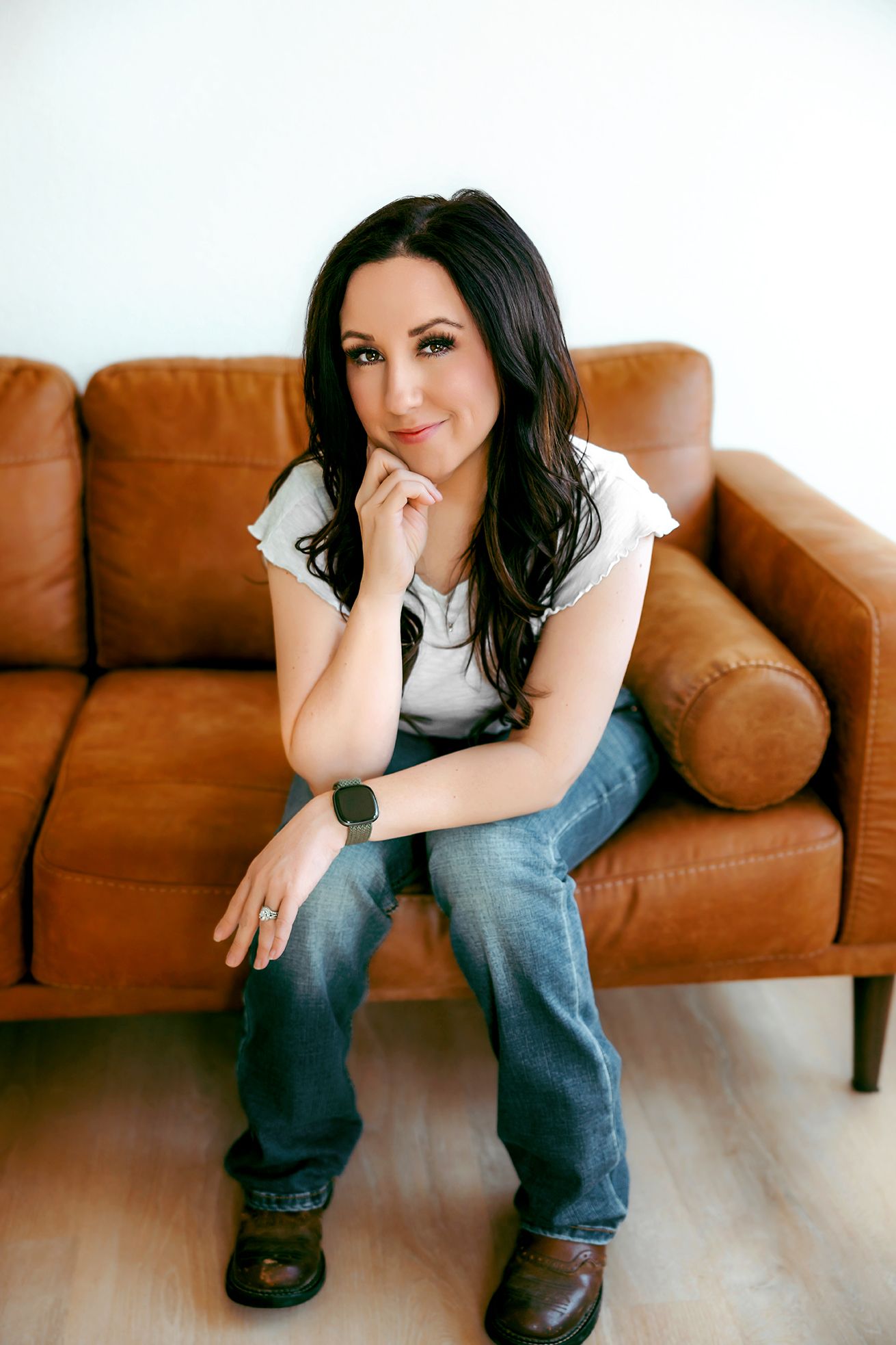 Casual portrait of someone relaxing on a brown leather couch wearing jeans and a white t-shirt in a neutral setting.