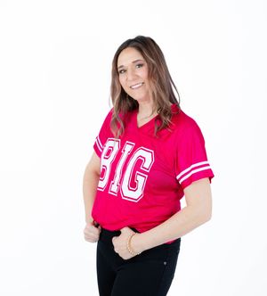 Person wearing a bright red collegiate-style t-shirt with "BIG" text and black pants poses against white background.