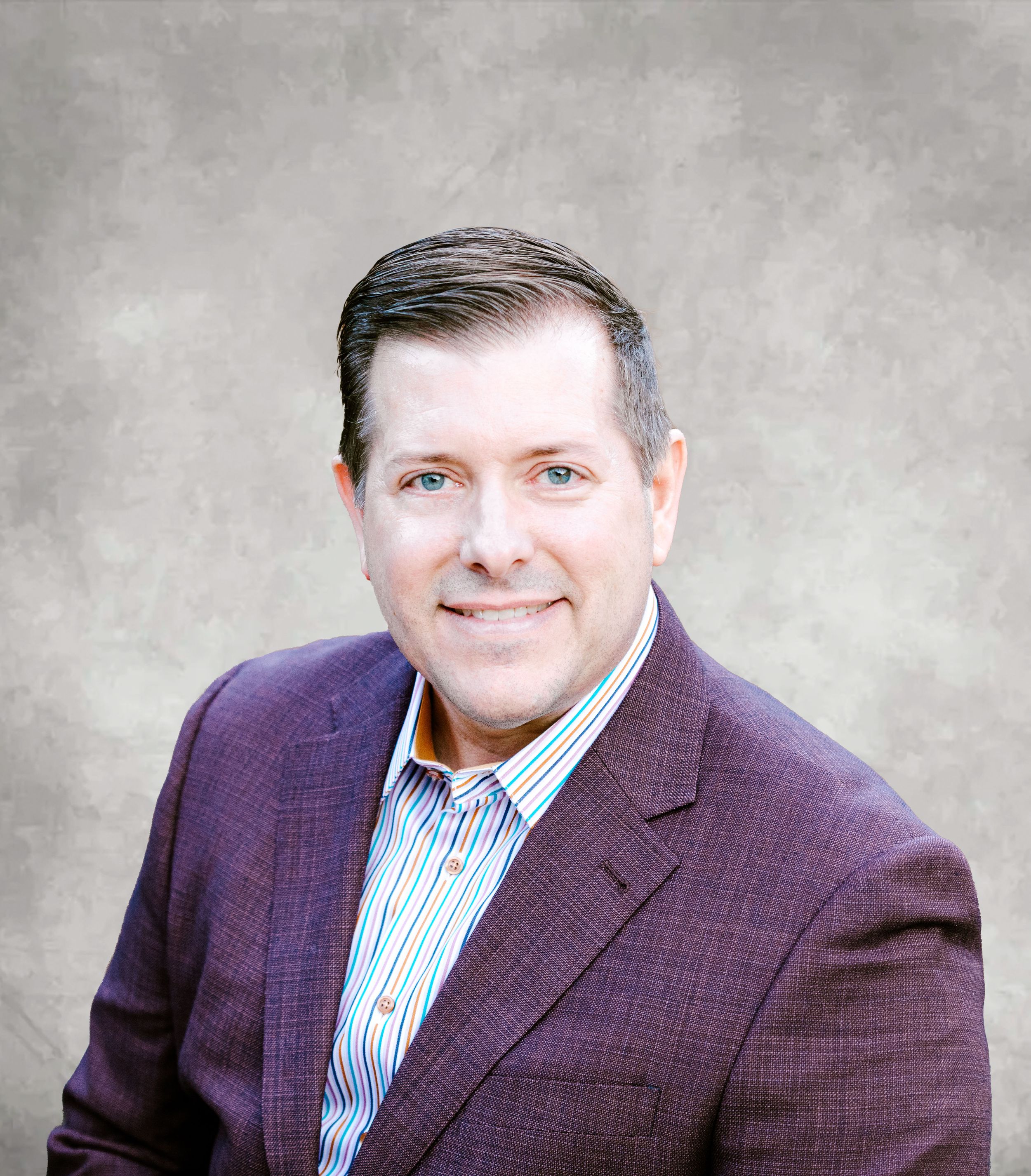 Professional headshot of person in purple blazer and striped shirt against grey background.