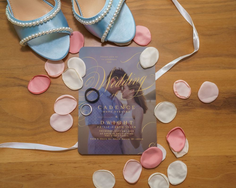 Wedding invitation with couple photo, blue heels, silver rings, and pink and white rose petals on wooden floor.