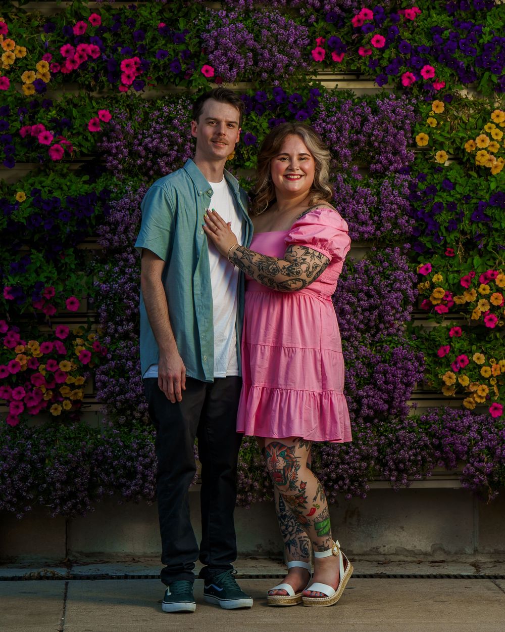 Two people stand against a vibrant floral wall with pink and purple blooms in a romantic outdoor setting.
