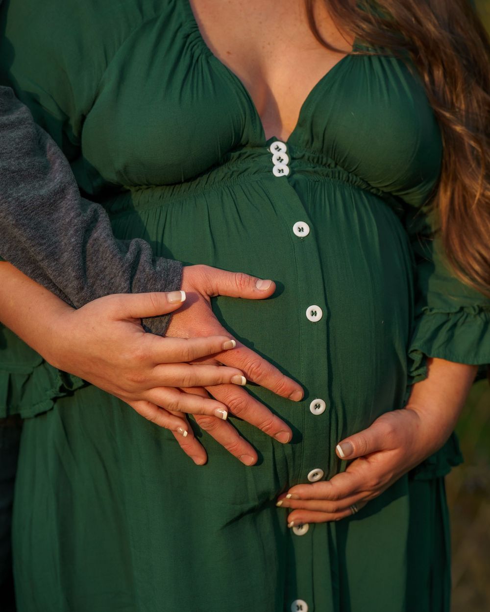 Close-up shot of hands resting on a pregnant belly in an emerald green button-down maternity dress.