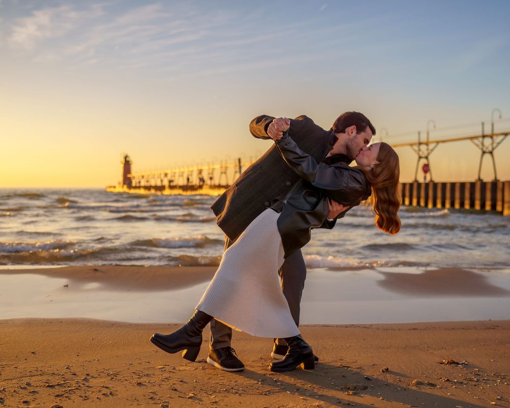 Couple sharing a romantic dip kiss on a sandy beach at sunset with a lighthouse in the background.