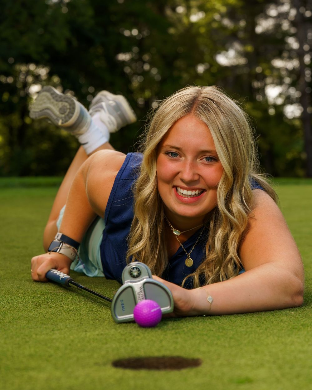Young blonde woman lying on a golf green, smiling, holding a putter near a purple golf ball beside the hole.