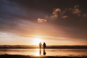 Two silhouettes hold hands while standing on a beach at sunset with golden light reflecting on the wet sand.