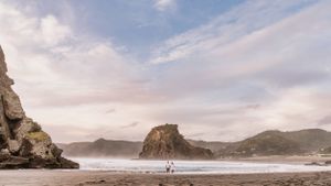 Two people walking on scenic Piha Beach with Lion Rock formation and coastal cliffs under cloudy sky in New Zealand.