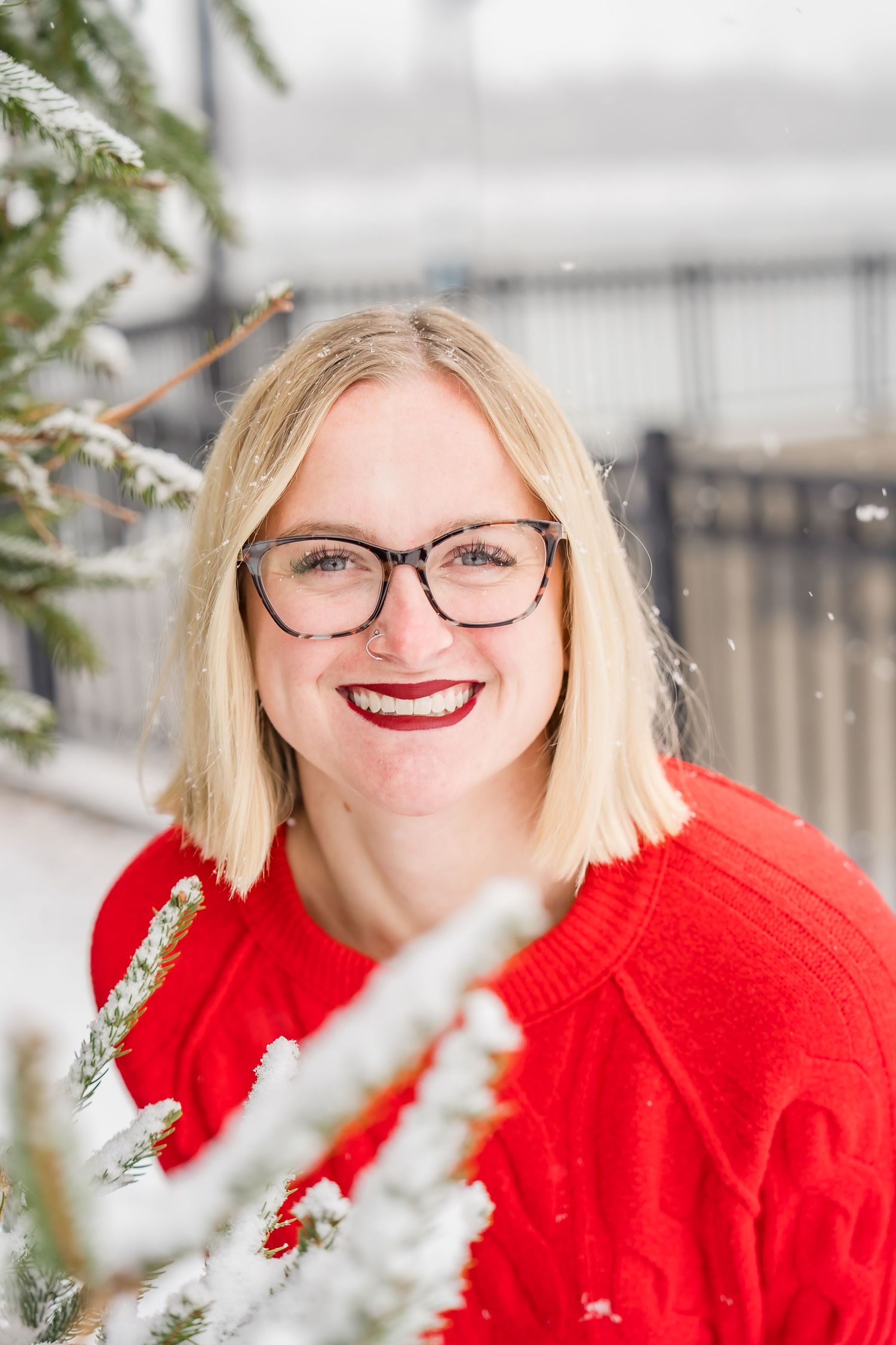 A person wearing glasses and a bright red sweater smiles while posing near snowy winter branches outdoors.