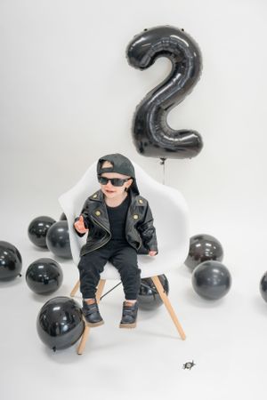 Stylish toddler in black outfit sits under number 2 balloon with black party decorations.