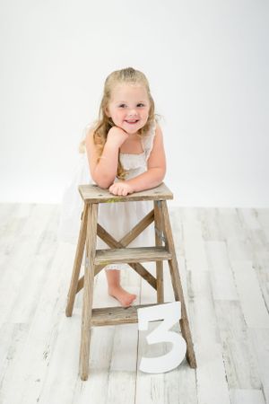 Small child in white dress sitting on vintage wooden stepladder with number 3.