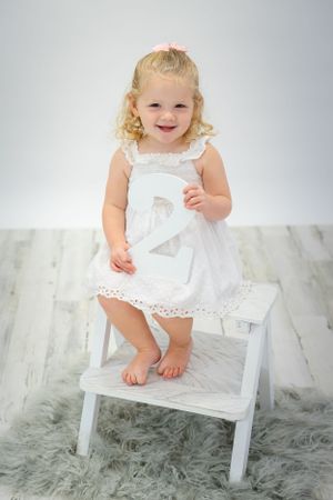 Small child in white lace dress standing on white steps with number 2.