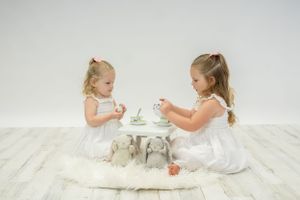 Two small children in white dresses having tea party on white floor.