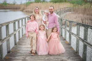 A family wearing coordinated pink and neutral outfits poses on a wooden boardwalk near wetlands.
