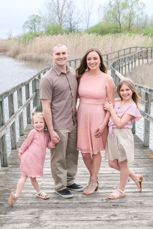 A family of four in coordinating pink and beige outfits poses on a wooden boardwalk by the water.