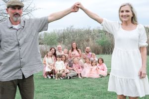 A couple holds hands while a group in pink dresses sits in the background near marsh grass.