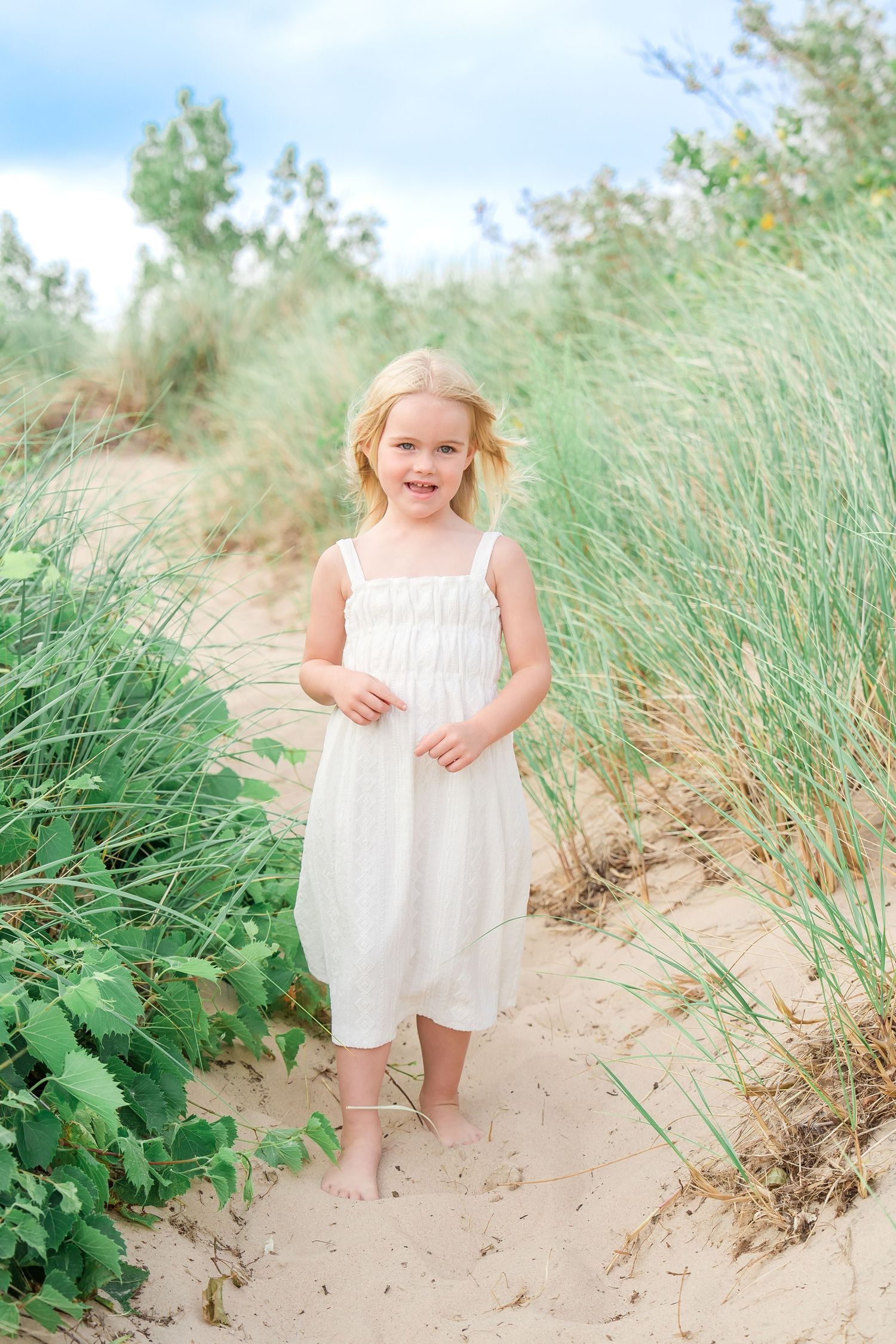 A child in a white summer dress stands barefoot on a sandy path surrounded by beach grass on a sunny day.