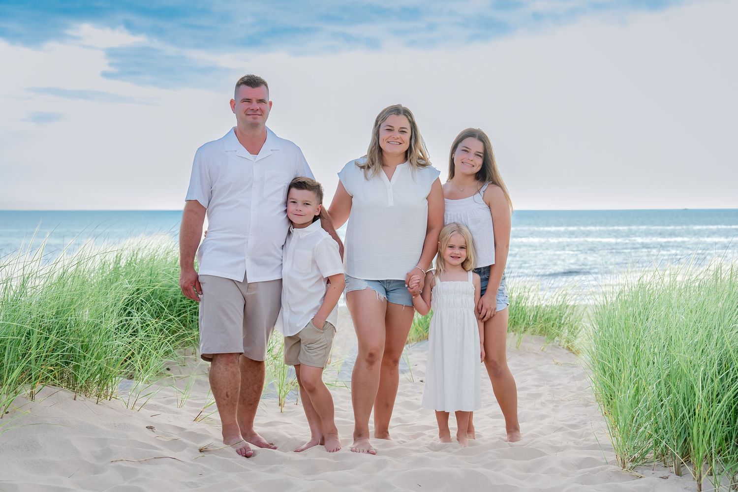 Family group wearing white and beige outfits poses together on sandy beach with ocean waves and sea grass in background.