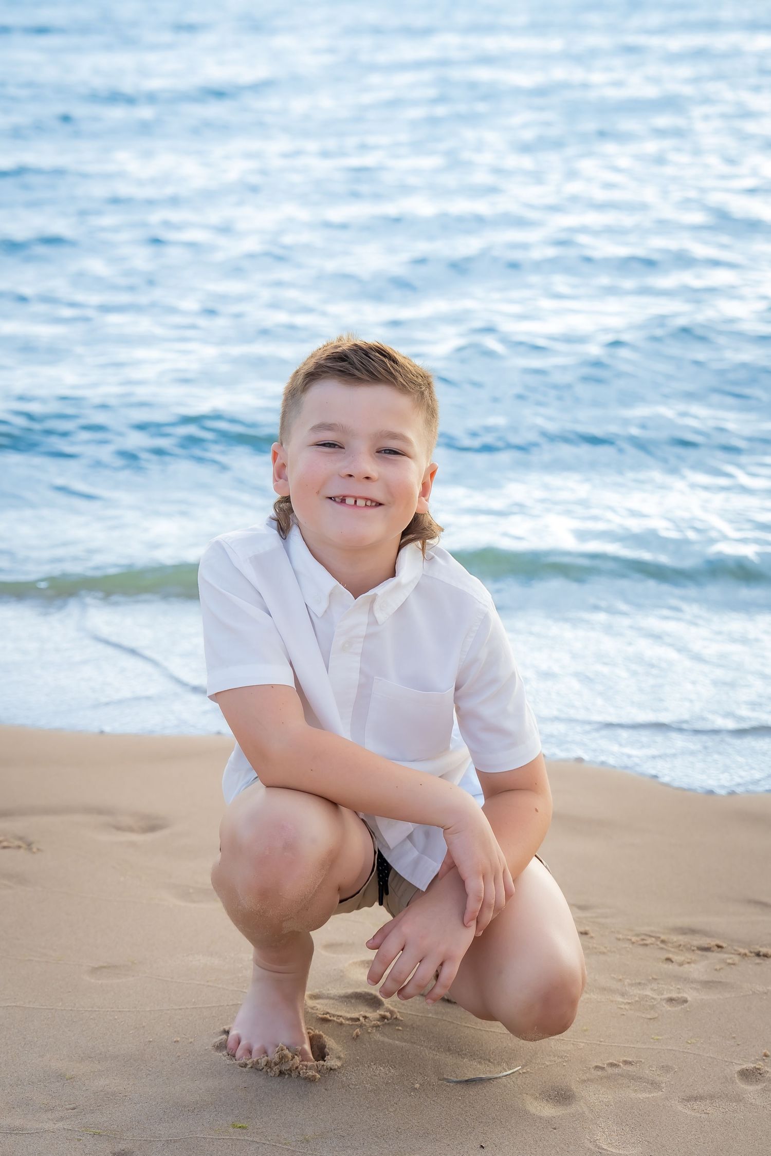 Child crouches on sandy beach wearing white shirt with ocean waves in background.