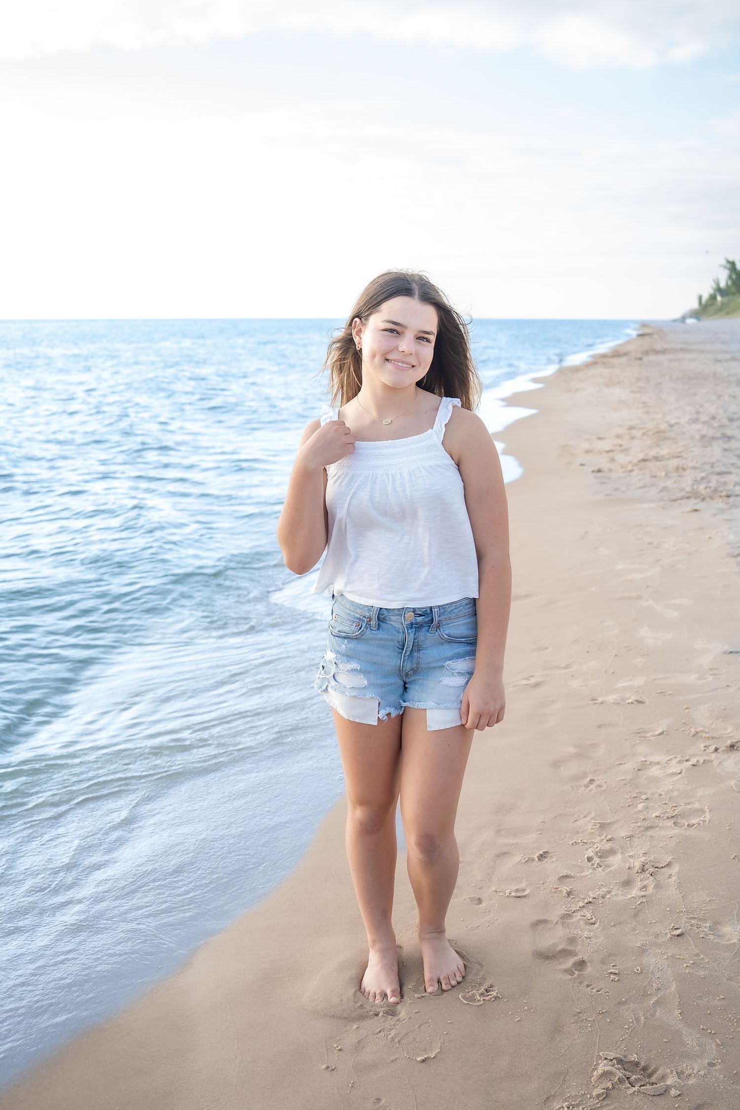 A person in a white top and denim shorts poses barefoot on a sandy beach with blue water and sky in the background.