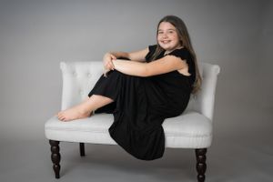 Portrait of person in black dress posing on white settee in studio setting.