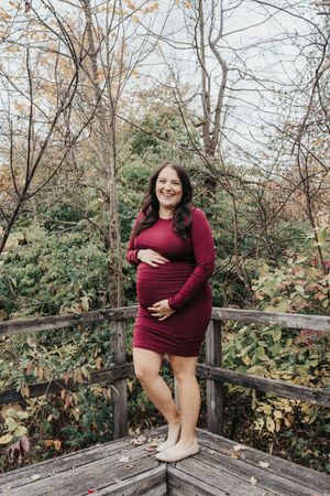 A maternity photoshoot on a wooden deck surrounded by autumn foliage features a woman in a burgundy dress.