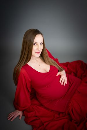 Woman in flowing red maternity dress poses against dark background during pregnancy photoshoot.