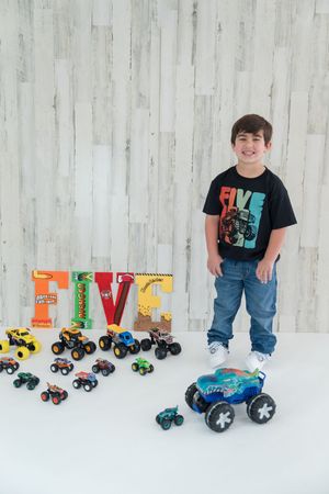 Colorful toy monster trucks and letter decorations displayed against white wall with young child in black t-shirt and jeans.