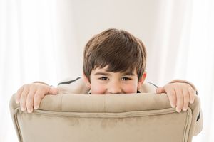 Two children peek playfully over the back of a beige couch in a living room setting.