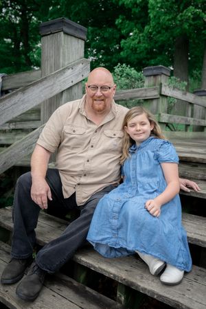 Two people in casual attire share a relaxed moment on wooden bleacher-style steps with green foliage in background.