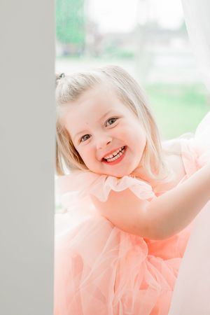 A small child wearing a peach-colored tulle dress smiles brightly in a soft, naturally lit setting.