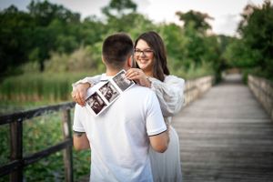 Couple on wooden bridge holding ultrasound photos during pregnancy announcement photoshoot.