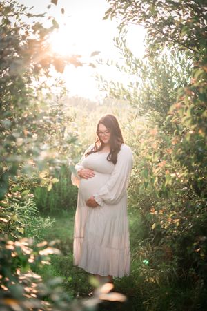 Ethereal maternity portrait in sunlit garden with flowing white dress.