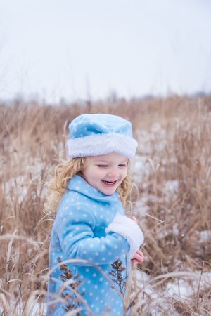 A joyful child in a light blue winter coat plays in a snowy field of tall dried grasses on a winter day.