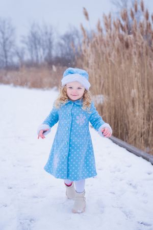 A small child wearing a blue polka dot winter coat plays in the snowy field with tall brown reeds in the background.
