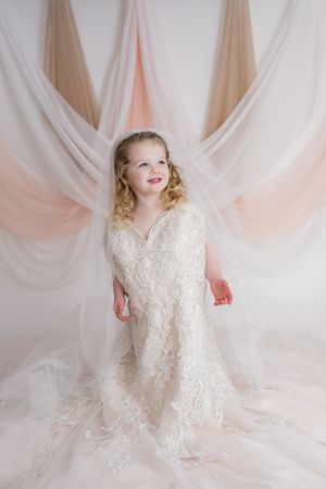 A child in a flowing white lace dress twirls gracefully against a soft pink draped backdrop.