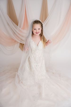 A young child in a flowing white lace dress twirls and dances against a soft pink and beige draped fabric backdrop.