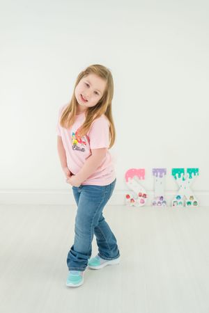 Young child in pink shirt and jeans poses playfully against white background with colorful decorations.