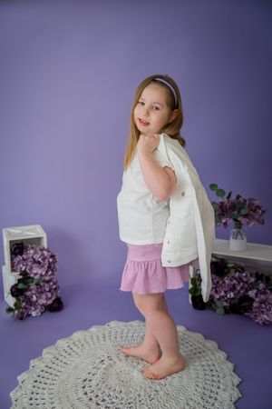 Young model in white top and pink skirt poses on crochet rug with purple hydrangeas and lilac wall background.