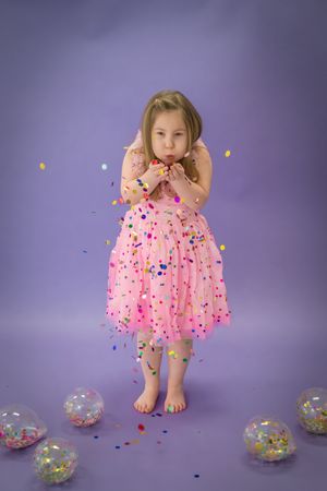 A child in a pink polka dot dress plays with colorful confetti and clear balloons against a purple backdrop.