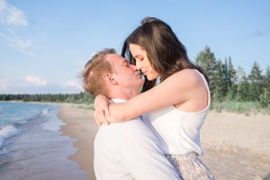 A couple shares a romantic embrace on a sandy beach with water and forest in the background.