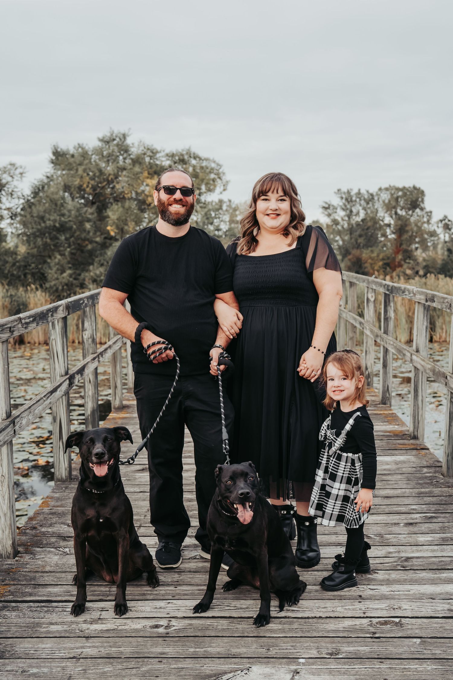 Family in black outfits poses with their black dogs on a wooden boardwalk during an outdoor photo session.