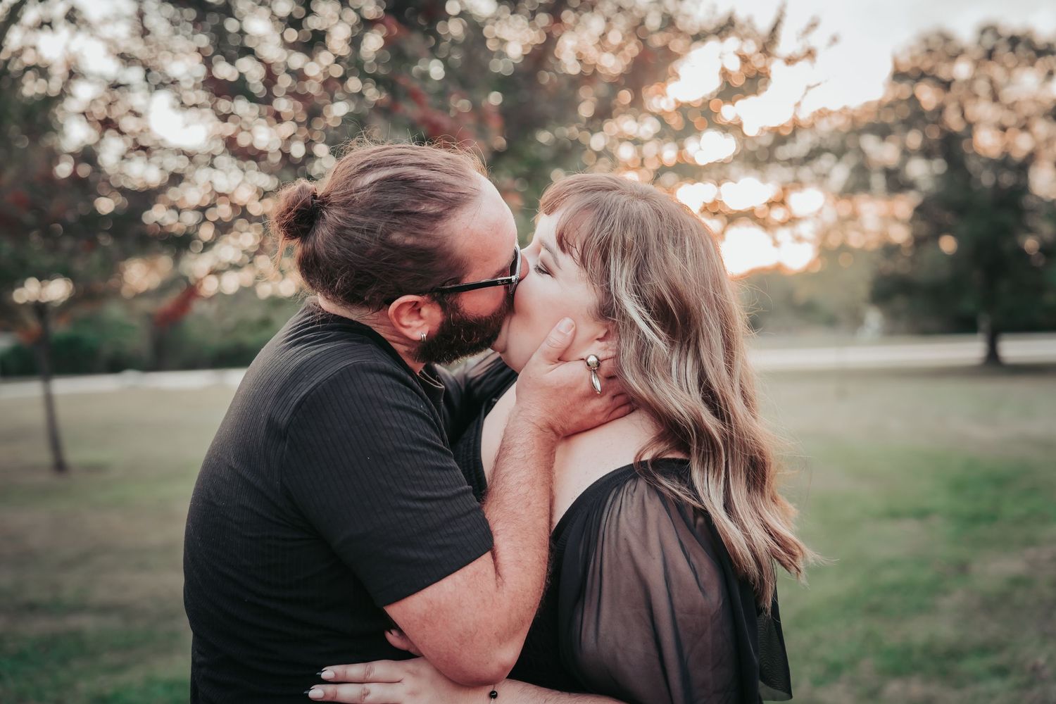 A couple shares a romantic kiss in a park during golden hour with sunlight filtering through trees in the background.