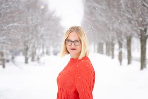 A figure in a bright red winter coat poses in a snowy forest landscape with snow-covered trees in the background.