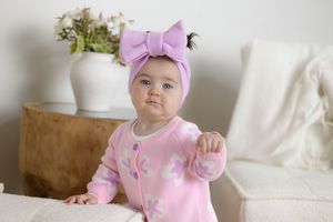 Adorable baby girl wearing a pink floral outfit and large lavender bow headband, sitting up and reaching forward.